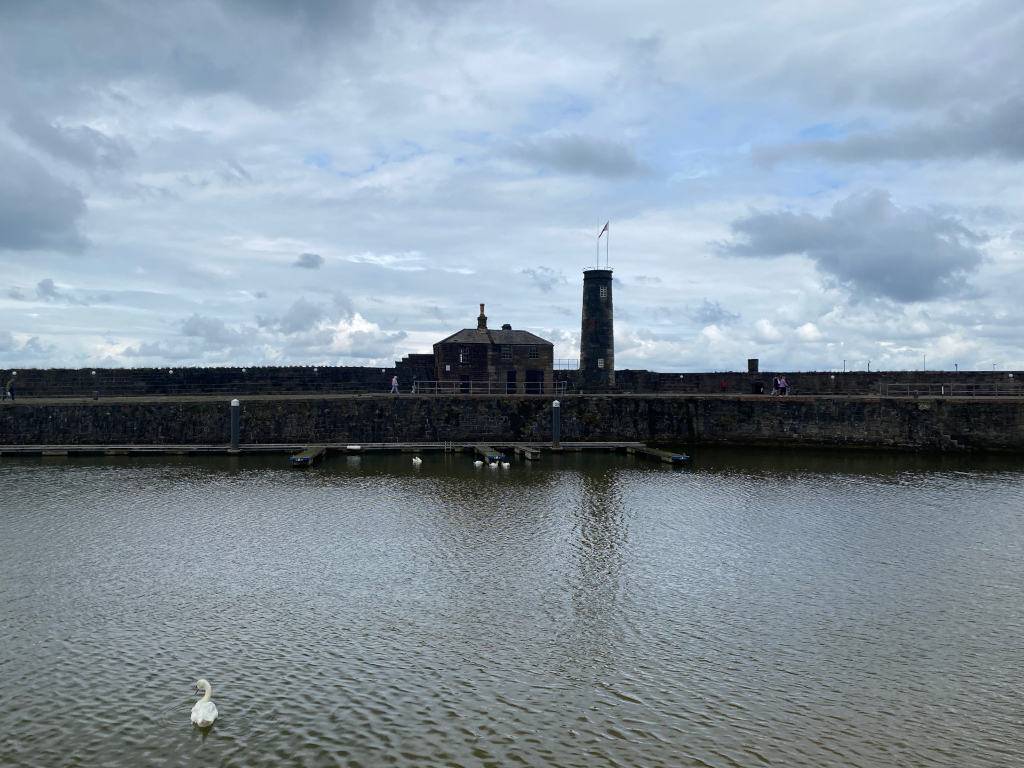 Calm body of water with a single swan swimming in the foreground.  In the background, a stone wall encloses a small marina with several moorings and a small building with a tall, slender tower. A few people are visible walking on the wall. The sky is overcast with dark, brooding clouds. The overall mood is serene yet somewhat sombre due to the weather.