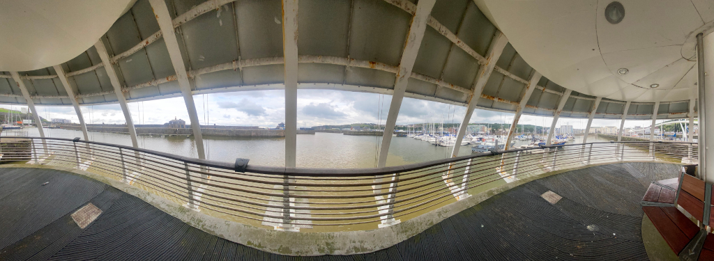 Panoramic view from a curved walkway or bridge overlooking a marina. The walkway has a modern, slightly weathered design with a metallic, grey and white structure. Through the railings, numerous boats are visible in the calm water of the marina, along with a waterfront area and buildings in the background. The overall scene suggests a peaceful harbor setting, possibly somewhere in a coastal town or city. The sky is mostly cloudy.