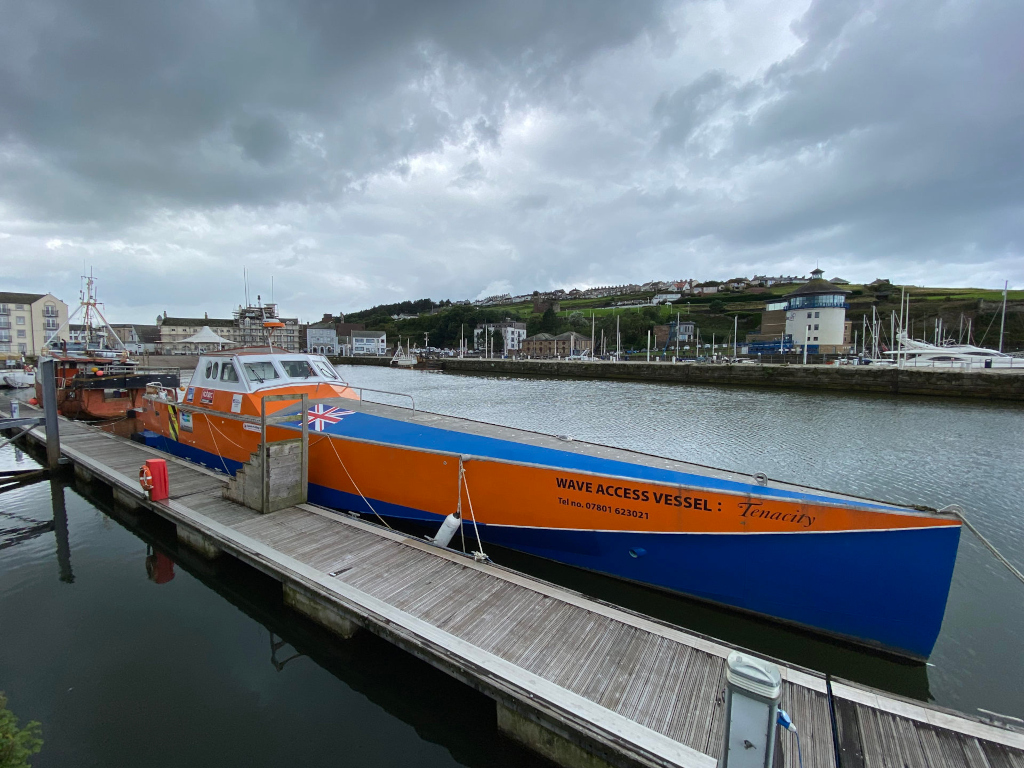 Vibrant orange and blue boat named Tenacity, identified as a Wave Access Vessel, docked at a marina. The boat is prominently featured, with its name and contact information clearly visible. The background includes a cloudy sky, a row of buildings, and other boats in the marina.