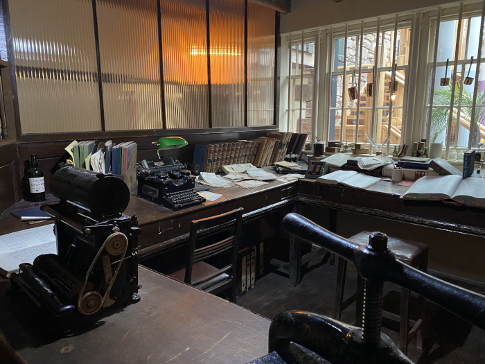 Dimly lit, antique-style office or study. The main focus is a large, dark wood L-shaped desk cluttered with books, papers, and antique writing implements including a typewriter and what appears to be a printing press or similar machine. A window with barred panes looks out onto an external stairwell and some greenery. The overall atmosphere is one of historical, potentially literary or academic work.