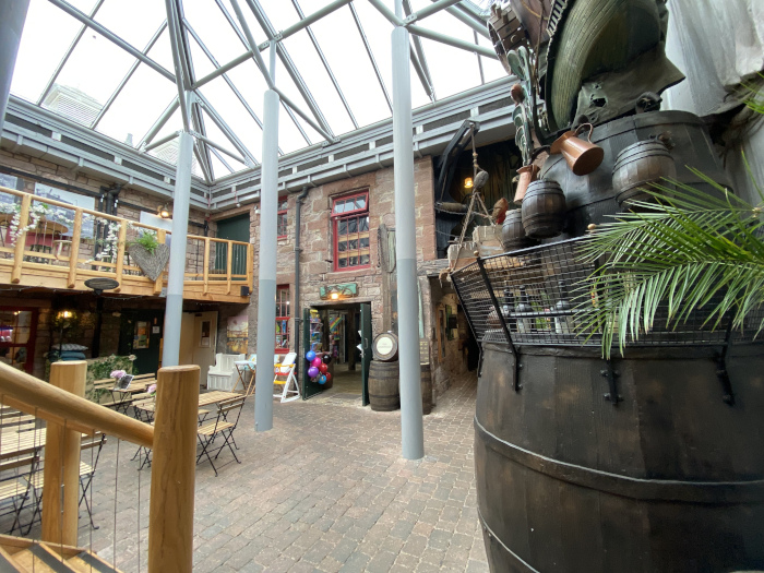 Interior courtyard of a building, possibly a shopping centre or marketplace. The courtyard has a glass ceiling, creating a bright, airy atmosphere. There are several shops or stalls visible, with one featuring balloons suggesting a celebratory or child-friendly atmosphere. A large, decorative whiskey barrel, possibly part of an art installation, dominates the right side of the image. The overall style is eclectic, blending modern architecture with elements of a nautical or pirate theme, as indicated by the barrel and possibly the ship-like structure above it. Wooden tables and chairs are arranged, suggesting seating for patrons. The architectural style also seems to incorporate elements from a historic building.