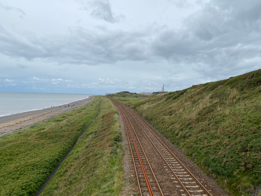 Coastal railway track running parallel to a beach.  The track extends into the distance, where a power plant or industrial facility is visible on the horizon. The foreground is characterized by grassy dunes and vegetation bordering the tracks and the beach. A calm sea is visible in the background under a cloudy sky. A small group of people can be seen on the beach in the distance. The overall atmosphere is somewhat bleak and quiet.