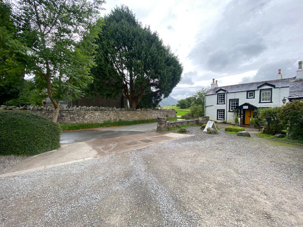 Gravel parking area in front of a white two-story building, which appears to be a pub or inn. To the left, there's a stone wall running along a narrow road, lined with lush green vegetation, including trees and hedges. The background includes a slightly overcast sky and hints of a distant mountain or hill. The overall feel is quaint and rural.