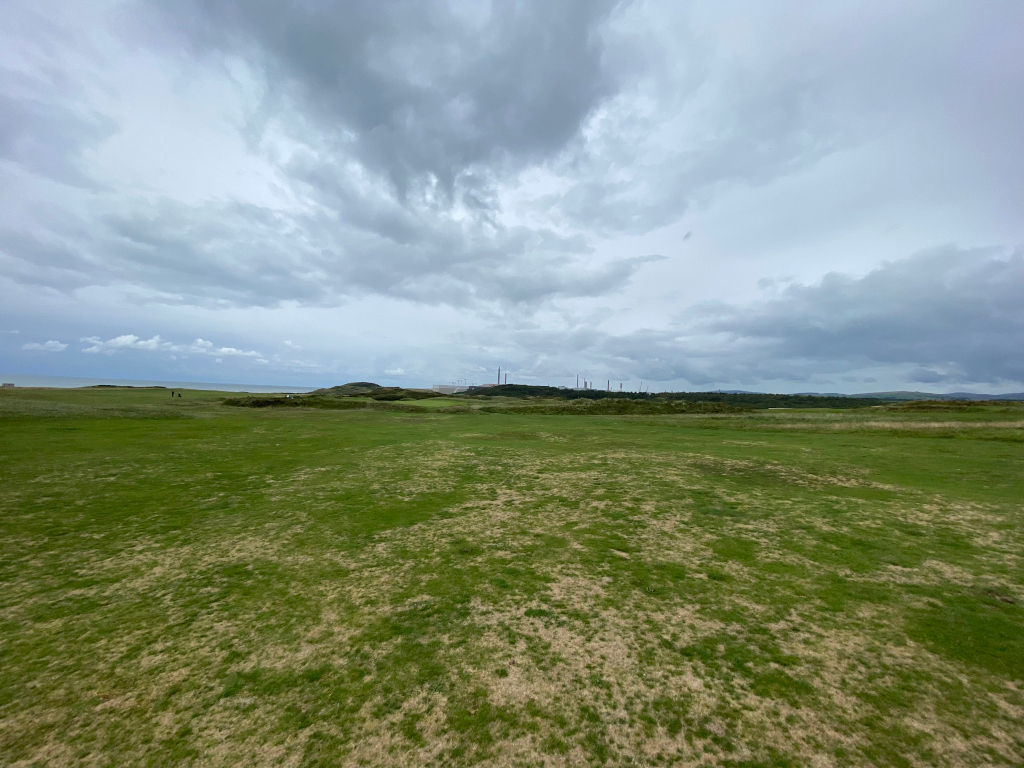 Wide, expansive view of a golf course fairway under a cloudy sky. The fairway is primarily green, with patches of brown or tan indicating possible dryness or dormancy. In the distance, a low-lying hill or dune is visible, beyond which industrial structures – possibly power plant cooling towers – are partially discernible against the overcast sky. A hint of ocean is visible on the far left horizon. The overall mood is somewhat somber or quiet, due to the overcast sky and the muted colors.