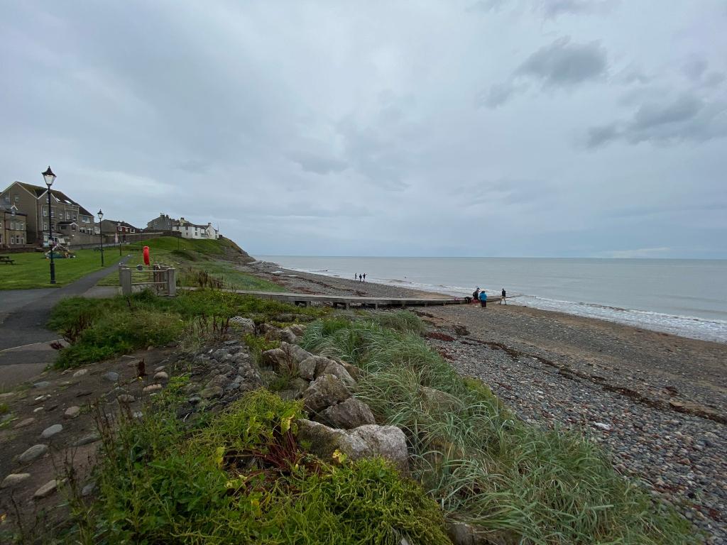 Coastal scene on an overcast day. In the foreground is a rocky, pebbly beach with vegetation growing along the edge. A paved walkway leads from a small residential area, seen in the background, down towards the beach. A few people are visible on the beach in the mid-ground. The overall mood is calm and somewhat subdued due to the muted colors and cloudy sky.