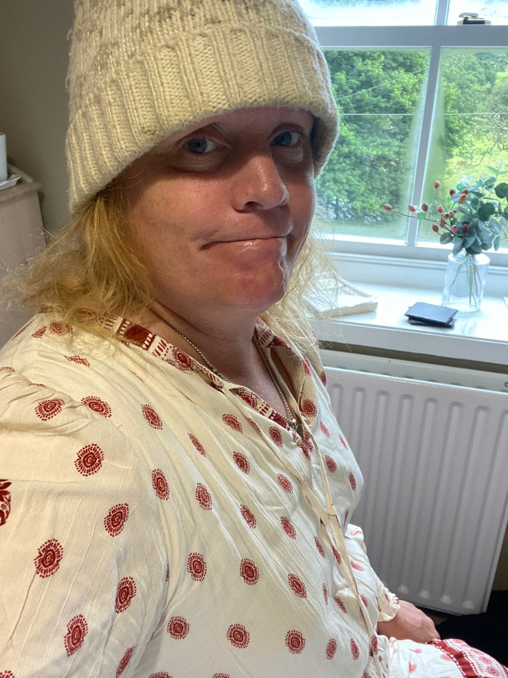 Close-up selfie of Leonie with shoulder-length blonde hair, wearing a cream-coloured knit beanie pulled low over their forehead, partially obscuring their eyes. She is also wearing a loose-fitting, off-white tunic or dress with a small, repeating red pattern. The background features a window with greenery visible outside and a radiator. The overall impression is of a casual, somewhat pensive portrait, taken indoors. Leonie's expression is neutral or slightly sombre.