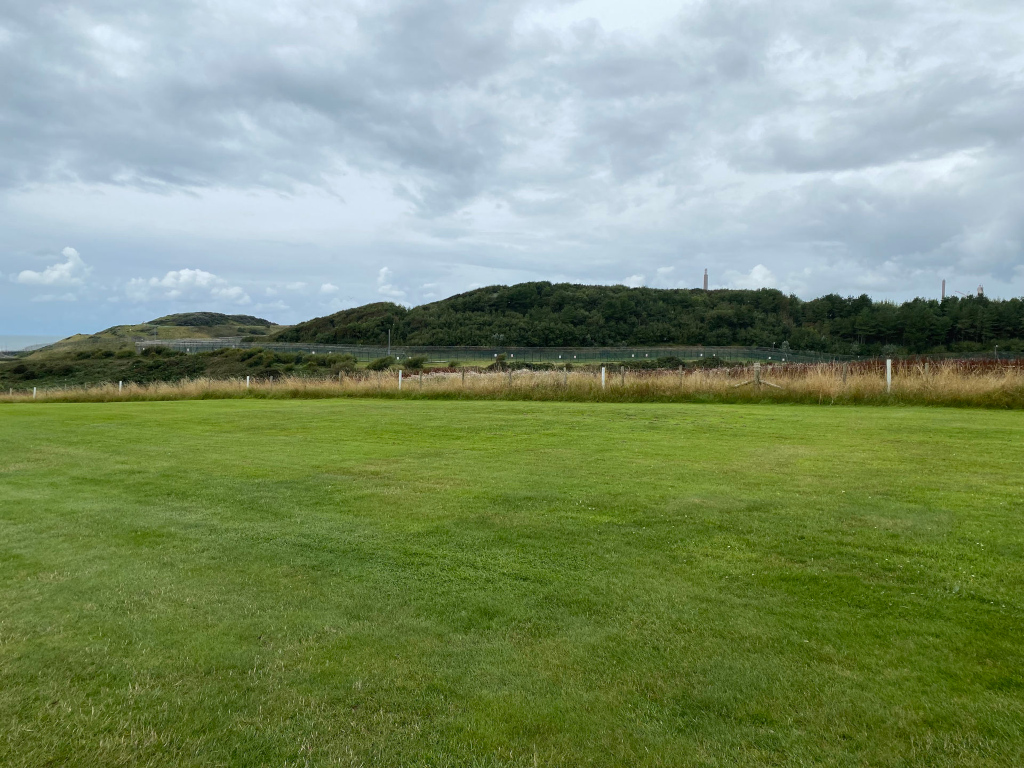 Grassy field in the foreground, stretching towards a line of tall grass and a fence. Beyond the fence are rolling green hills covered with trees. In the far distance, atop the hills, there are two tall, slender structures, possibly towers or chimneys.