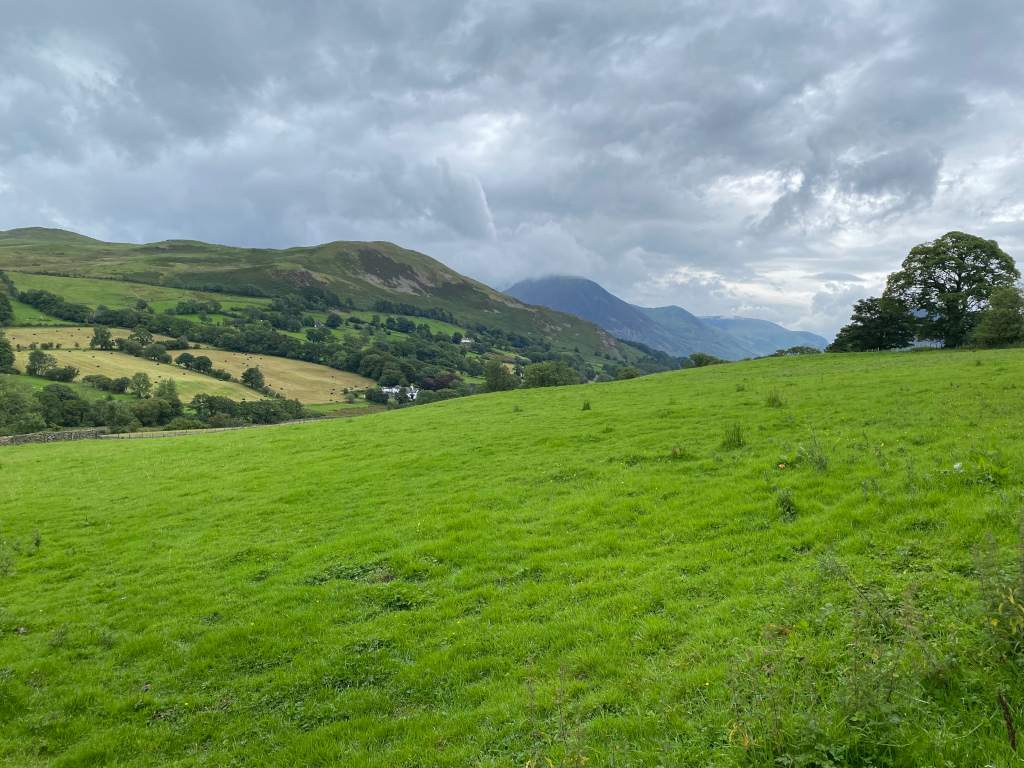 Lush green field in the foreground, gently sloping upward towards a valley. Beyond the field, rolling hills and mountains stretch into the distance under a cloudy sky. Hay bales are visible in the mid-ground, suggesting agricultural activity. A few trees are scattered across the landscape. The overall impression is of a serene and pastoral scene in a mountainous region.