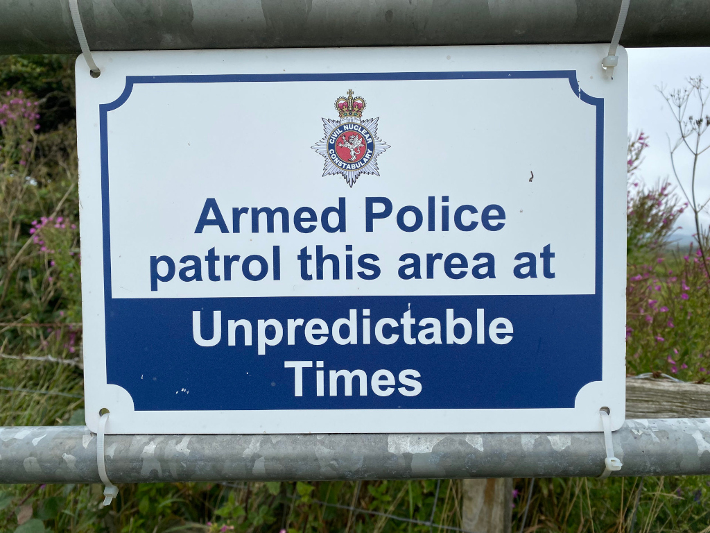 Sign stating that armed police patrol the area at unpredictable times. The sign features the logo of the Civil Nuclear Constabulary. It's attached to a metal fence post in what appears to be a rural or semi-rural setting.