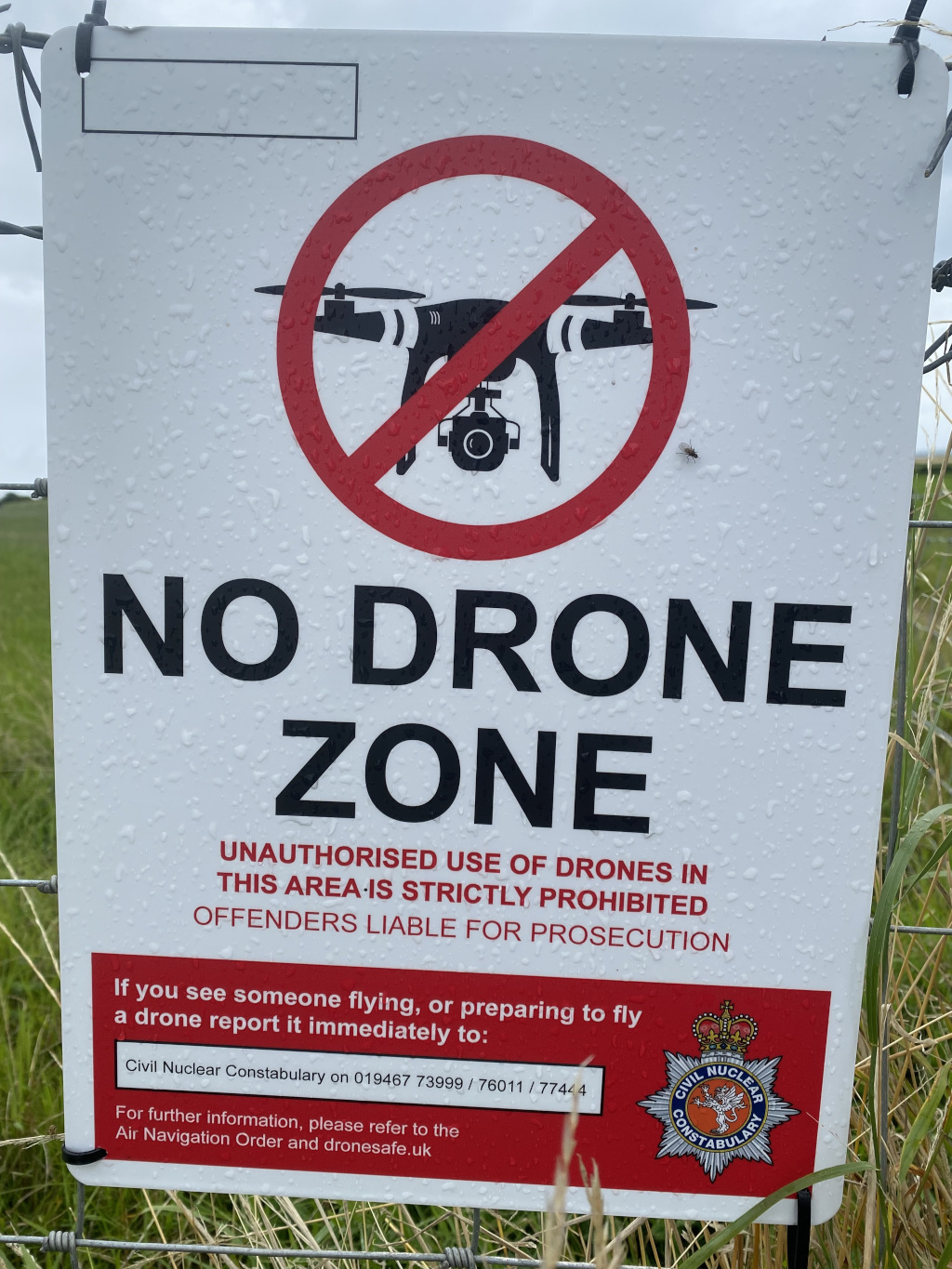 Sign prohibiting the use of drones in a specific area. The sign, which appears to be affixed to a fence, features a large red circle with a diagonal slash across an image of a drone, clearly indicating that drone use is forbidden. The text emphasises the strict prohibition, warns of prosecution for offenders, and provides contact information for reporting violations to the Civil Nuclear Constabulary. The overall message is one of security and restriction, likely intended for a location sensitive to unauthorised aerial surveillance. The slightly blurry background suggests an outdoor, possibly rural, setting.