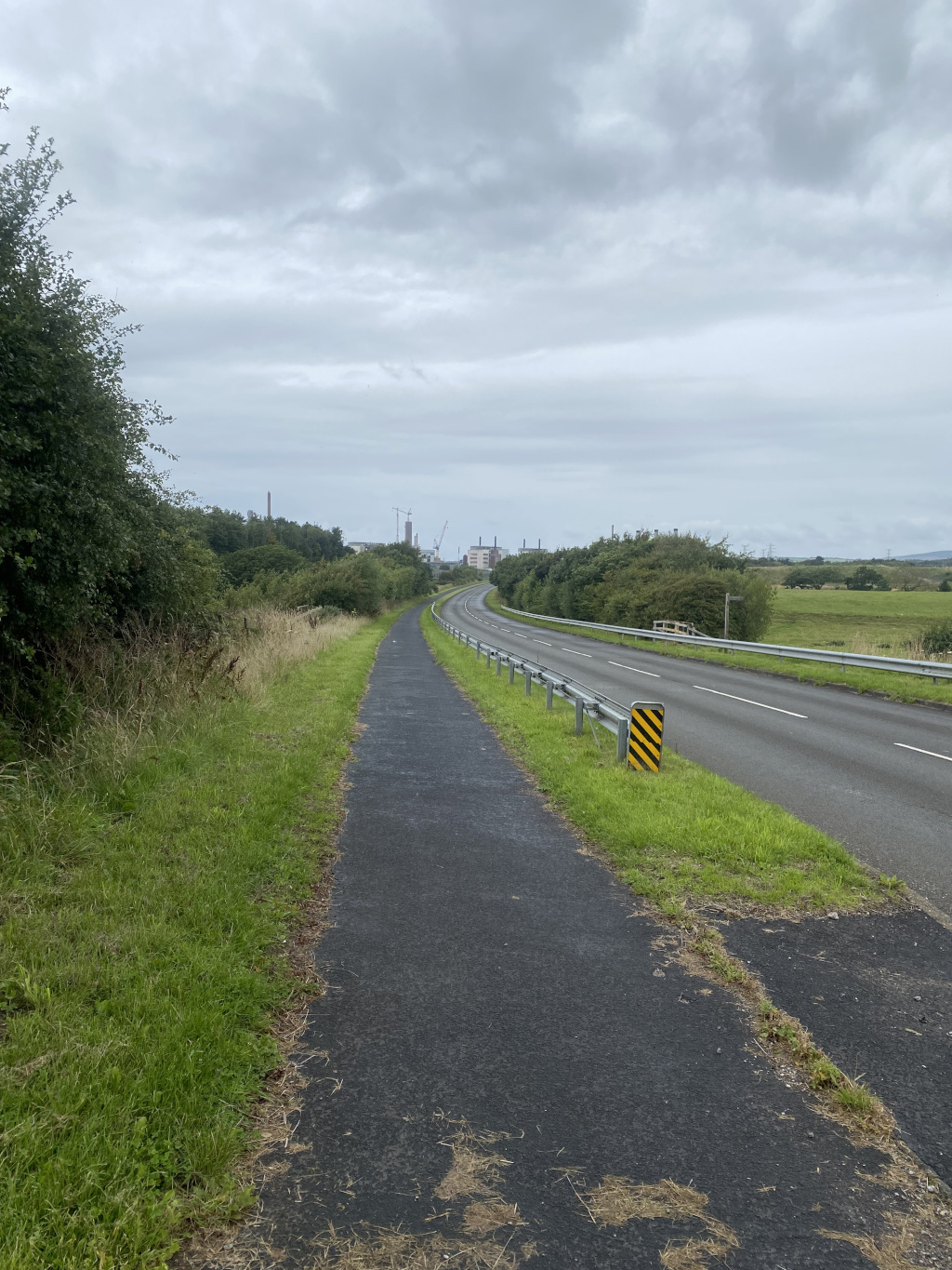 Paved path running alongside a two-lane road. The path and road curve gently to the right. To the left of the path is uncut grass and some bushes. On the other side of the road, more grass and some trees are visible. In the far distance, industrial structures, possibly a factory or power plant, can be seen under a cloudy sky. The overall mood is muted and somewhat industrial.