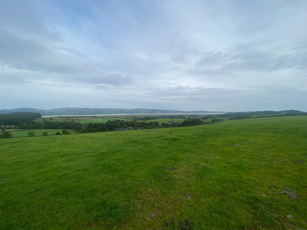Panoramic view of a verdant landscape. In the foreground, a vast expanse of lush green pasture dominates the scene. Beyond the pasture, the land gently rolls into a valley, revealing a tranquil body of water, possibly a river or estuary, nestled between low-lying hills. The sky is mostly cloudy, lending a somewhat muted but peaceful ambiance to the overall scene. 