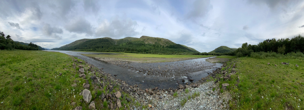 Panoramic view of a serene landscape. A calm river, with a rocky bed, flows through a verdant valley, flanked by gently sloping hills covered in lush green vegetation. The sky is mostly cloudy, adding a soft, diffused light to the scene. 