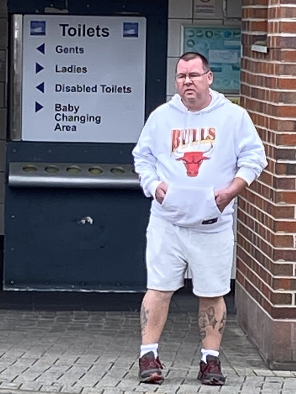 Middle-aged man with short, light brown hair and glasses standing in front of a public restroom sign. He is wearing a white hoodie with a Chicago Bulls logo, white shorts, white socks, and dark-coloured trainers. He has tattoos visible on his lower legs. The man appears to be waiting or standing idly. The overall setting suggests a public space, possibly a train station or similar location.