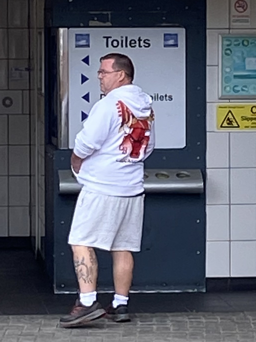 Middle-aged man, possibly in his 40s or 50s, standing in front of a public restroom. He's wearing a white hoodie with a large, stylised bull image on the back, white shorts, and dark-colored sneakers. His legs show a visible tattoo. The overall setting appears to be a public space, perhaps a train station or similar transit facility, based on the signage. The man seems to be waiting or pausing, with a somewhat neutral expression.