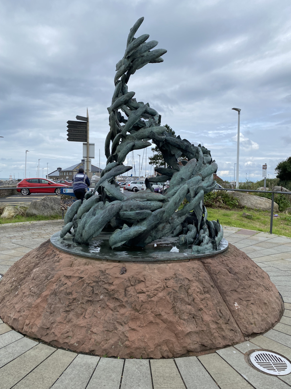 Bronze sculpture of a school of fish, dynamically arranged as if leaping or swimming. The sculpture sits atop a large, rounded, reddish-brown stone base. The background includes a parking lot, some buildings, and a partly cloudy sky. A person in a dark jacket is visible in the distance, near a directional signpost. The overall scene suggests a coastal or waterfront location.