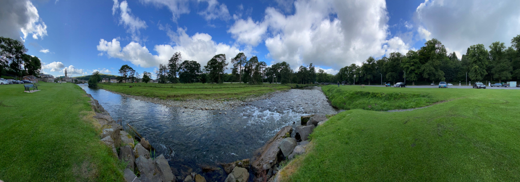 Panoramic photograph depicts a tranquil river scene. The river, relatively shallow and clear, flows gently through a grassy landscape. Trees line the riverbanks, creating a natural frame. A parking lot with several cars is visible in the background on the right, suggesting a public area. A small park-like area with a bench is seen on the left.  The overall impression is one of serenity and natural beauty. The sky is bright, partly sunny with fluffy clouds.
