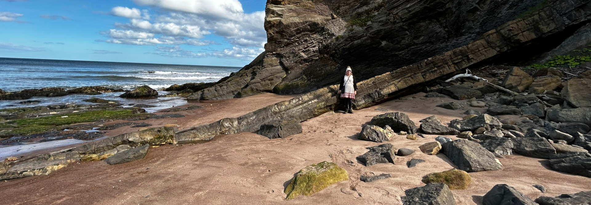 Leonie  standing on a sandy beach next to a large rock formation. The rock formation is dark and layered, and appears to have been shaped by erosion. The ocean is visible in the background, with waves breaking on the shore. The overall mood is peaceful and serene.