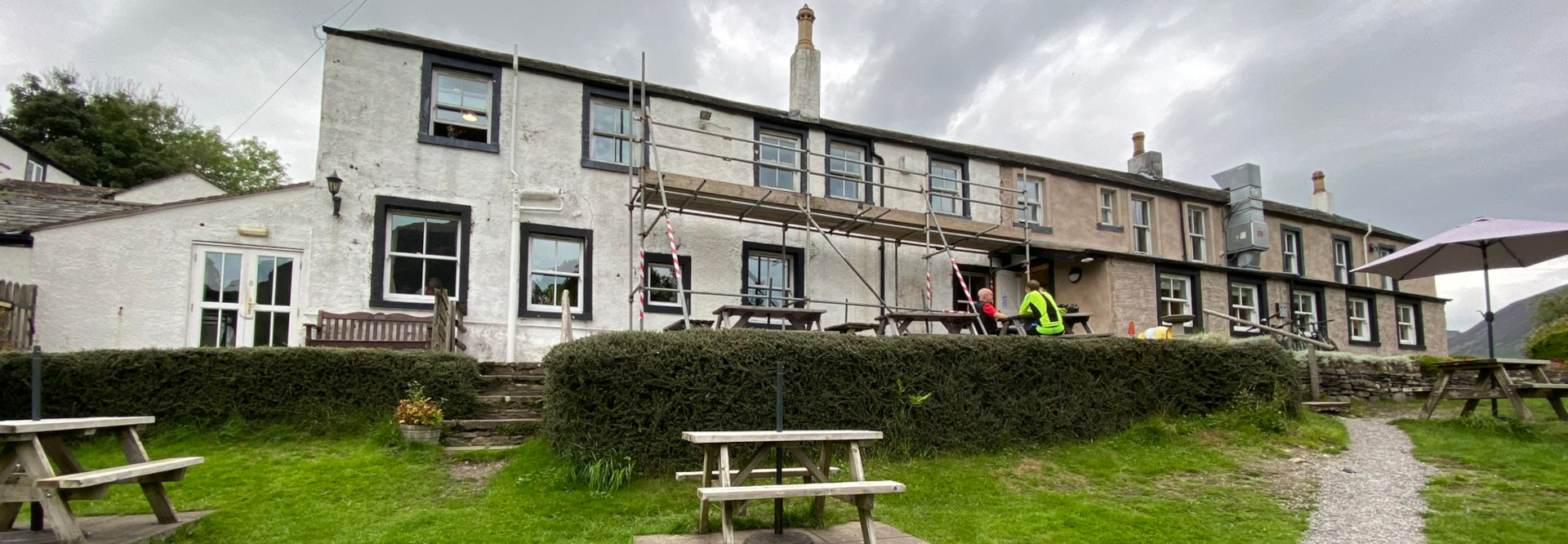 Long, two-story building, which appears to be a pub or inn, undergoing renovations. Scaffolding covers a portion of the building’s facade. Two people are seated at a picnic table outside, seemingly taking a break from the work or enjoying the view. The building has a rustic, traditional style, with stone and whitewashed walls, dark window frames, and several chimneys.  The setting is rural, with grassy areas, hedges, and picnic tables in the foreground. A portion of the building shows a different color of plastering, suggesting the renovations are underway. A parasol stands on the lawn.