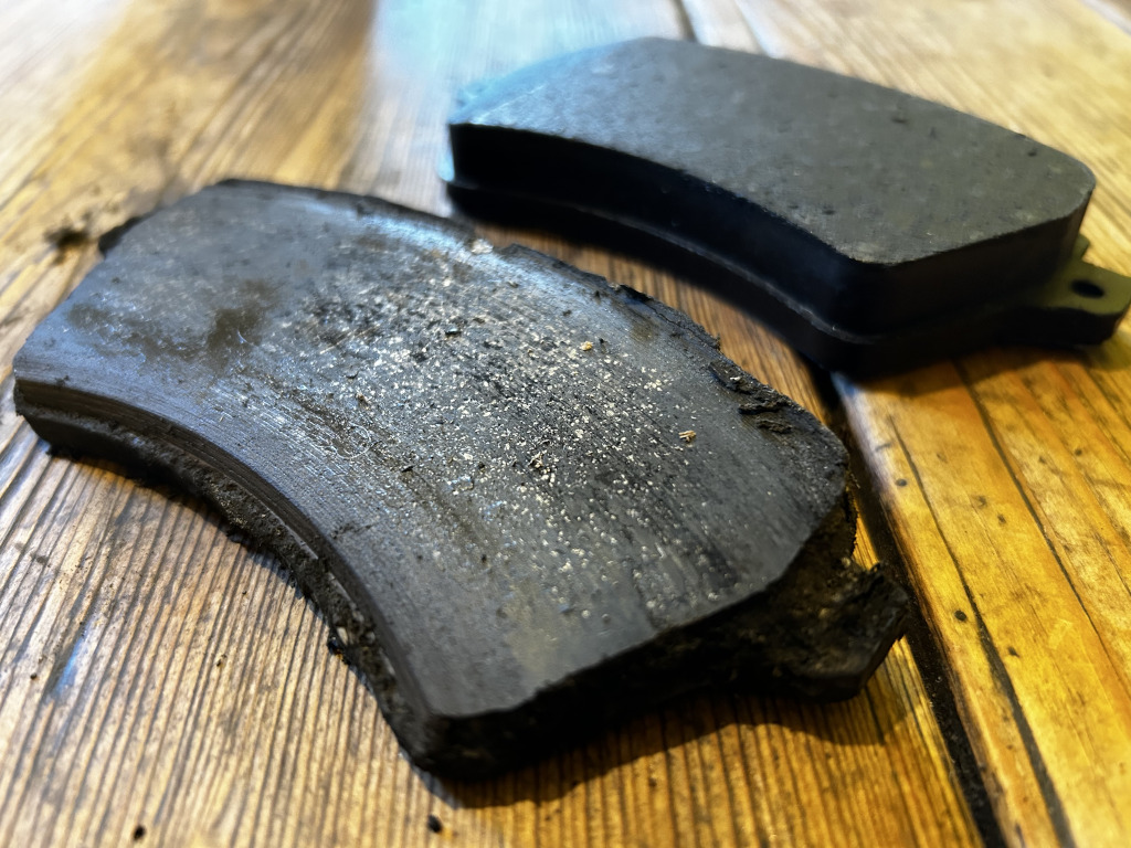 Two worn-out brake pads resting on a wooden surface. One brake pad (the upper one) is relatively intact, exhibiting a rectangular shape with slightly rounded edges. Its surface is dark charcoal grey-black, showing a matte texture with subtle, uneven wear. The other brake pad (lower) is significantly damaged and fragmented. It's cracked and broken along one side, revealing a ragged, irregular edge. Its surface shares the same dark grey-black hue as the other pad, but displays heavier wear, showing a dusting of light grey-white residue likely from brake dust, and exhibiting more pronounced, almost flaky textures around the broken areas. Both pads appear to be made of a dense, compressed material. They lay at a slight angle on the wood, with the damaged pad slightly overlapping the less damaged one. There's no indication of any human presence.