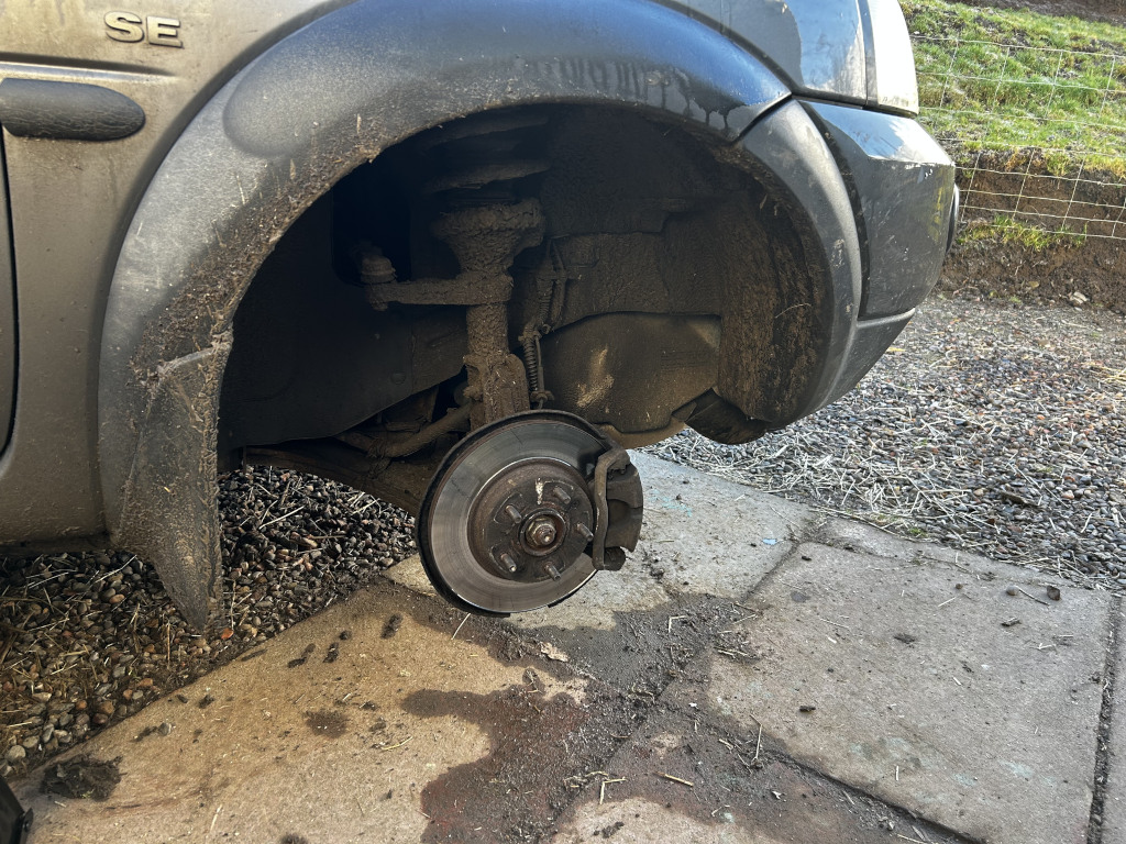 Close-up view of a lifted wheel of a dark gray Land Rover Freelander, model indicated by the SE badge subtly visible on the vehicle's fender.  The wheel is completely detached, showcasing the brake rotor and caliper assembly. The tire is absent. The vehicle's suspension components are clearly visible, coated in a layer of mud and dirt, suggesting recent off-road use or exposure to harsh weather. The wheel well is also heavily soiled, matching the overall dirty state of the visible parts of the vehicle. The ground beneath the car is a mixture of gravel, small stones, and patches of mud, suggesting an outdoor, possibly unpaved, setting.