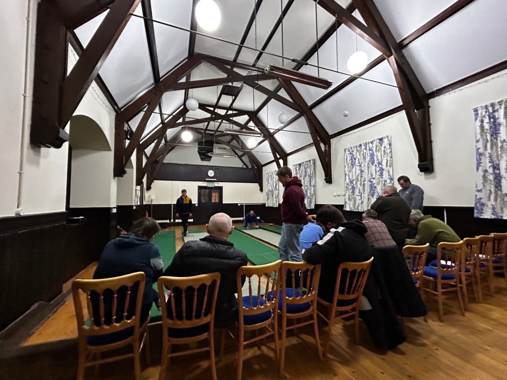 Group of men engaged in what appears to be a bowls game or similar indoor sport within a large hall. Two individuals are actively playing on a green matted bowling surface. One, wearing a dark jacket and possibly a yellow undershirt, appears to be throwing or preparing to throw a ball; the other, further down the lane, seems to be observing the trajectory. A row of men, mostly dressed in casual attire like jackets and sweaters in muted earth tones, sits on wooden chairs facing the playing area. Some appear engaged, watching the game intently, while others may be more casually observing. Their posture varies from attentive to slightly slumped, conveying different levels of engagement. The game takes place inside a high-ceilinged hall with exposed dark wooden beams forming a striking architectural feature. The ceiling appears high, light, and slightly arched, creating a sense of spaciousness.
