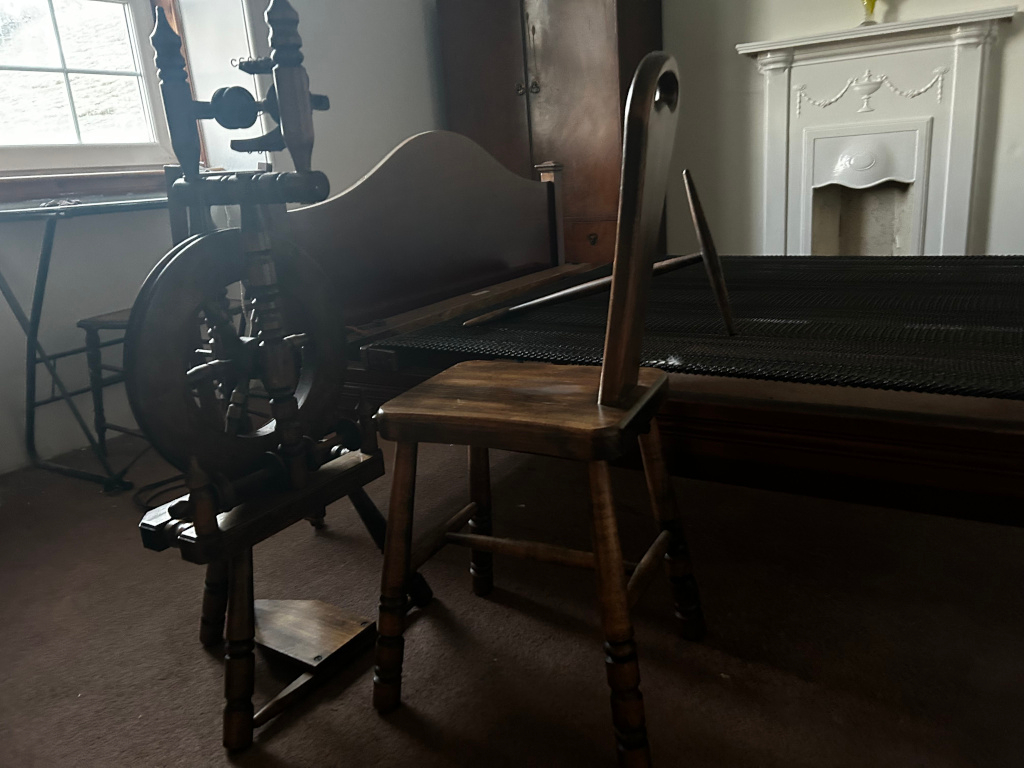 Dark brown, antique spinning wheel positioned slightly off-centre to the left.  Its wooden frame is intricately detailed, with small, turned spindles and a large, dark wood wheel.  The wheel appears to be in a resting state, not in active use.  To the right and slightly behind the spinning wheel sits a simple, rustic wooden chair. This chair, also dark brown, features a square seat and straight, slightly tapered legs, possessing a similar aesthetic to the spinning wheel, suggesting a harmonious, possibly historical setting. A dark brown wooden bed with a black mesh mattress occupies the background. A small wooden side table is partially visible near the left side of the image. The furniture is all dark, heavily shaded.