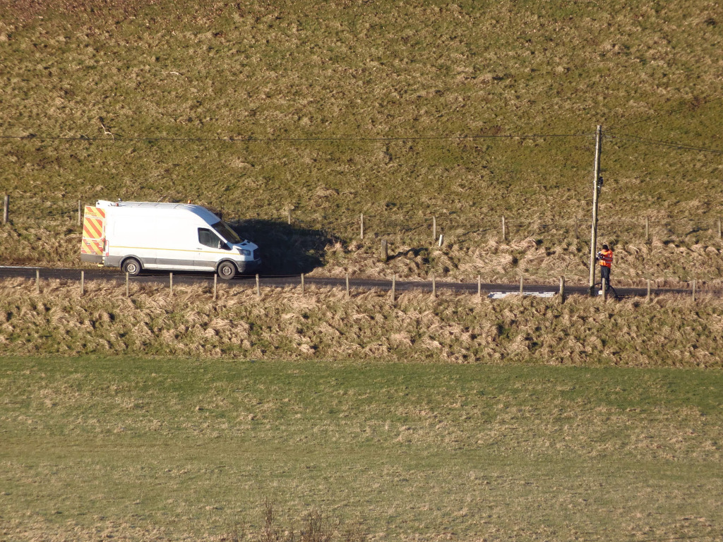 White panel van, slightly angled to the left, sits parked on the edge of a dark asphalt road. Its side is clearly visible, displaying diagonal orange and yellow reflective stripes on the back panel. The van appears to be a standard commercial vehicle, neither overly new nor particularly old. A person, small in scale due to distance, stands near a wooden utility pole on the right side of the road. They are wearing an orange-red vest or jacket, suggesting a worker's uniform. Their posture suggests they are working on or inspecting something near the pole. The scene is set in a rural environment, characterised by rolling, grassy hills that form the dominant background. The grass is predominantly a dry, brownish-green, suggesting it might be late autumn or early spring. Patches of lighter, almost straw-coloured dried grass are visible, particularly along the road's edge. A wire fence runs alongside the road.