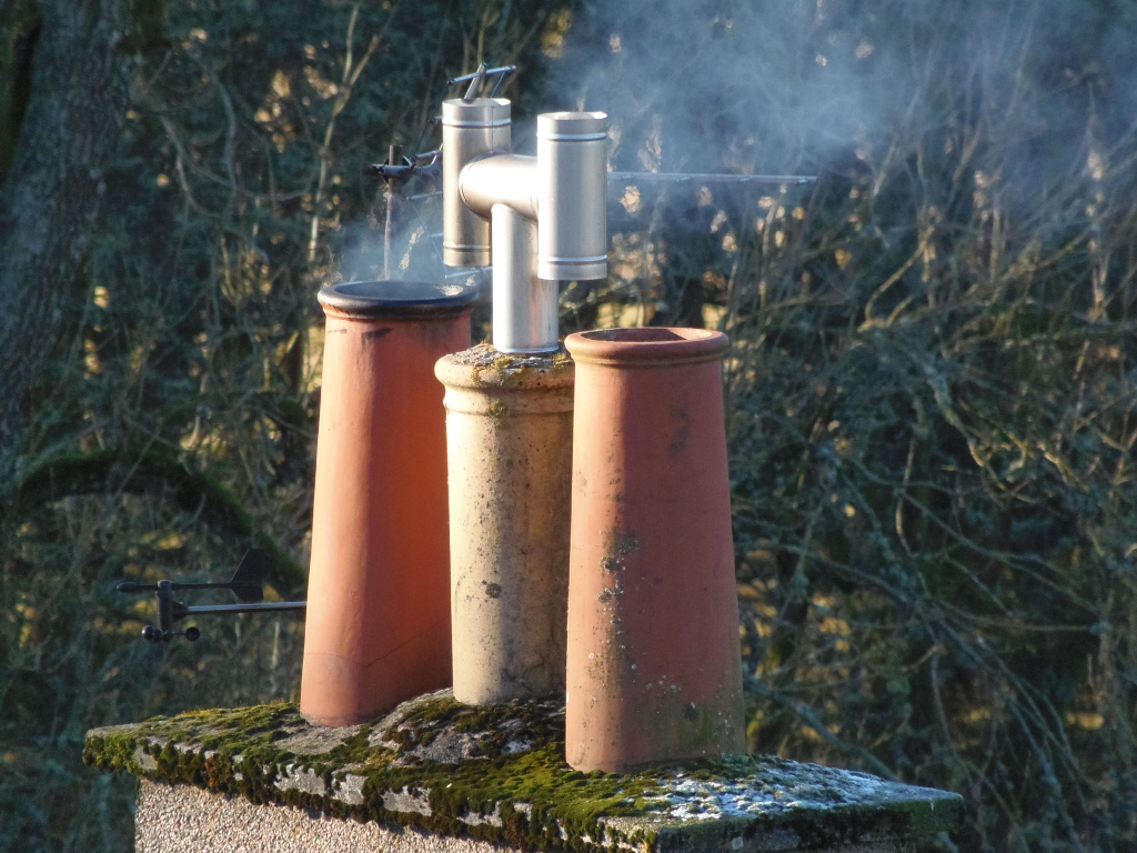 Chimney assembly atop a moss-covered stone structure. Three terracotta chimney pots, varying slightly in shade and exhibiting a subtle texture of age and weathering (moss and small discolourations), are arranged closely together. The pots are cylindrical, slightly tapered, and of a muted reddish-brown hue. A modern, metallic, stainless steel-appearing flue system is positioned centrally above the pots. This flue has a slightly angled, double-pipe design, suggesting efficient venting. Smoke, a pale greyish-white, softly rises from the top of the flue, hinting at a gentle fire within. A small, dark-coloured weather vane, with simple arrow-like arms, is attached to the chimney base, slightly off-centre to the left.
