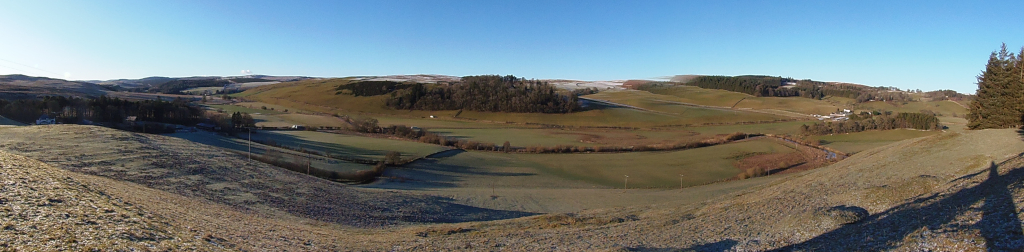 Panoramic landscape view devoid of human figures. The main objects are undulating hills and fields, predominantly grassy with patches of brown and hints of frost. A strip of dark-green woodland runs horizontally across the middle ground. There are a few small structures, perhaps farmhouses, barely discernible in the middle distance. The foreground is composed of a steep, gravelly hill, showing signs of frost or light snow. On the far right, a cluster of evergreen trees stands prominently. The scene is rural, likely in a temperate climate. The background consists of rolling hills extending to the horizon. Some distant hills show hints of a pale, light dusting of snow. The lighting indicates a bright, sunny day, with deep shadows cast by the sun angled at a relatively low position.