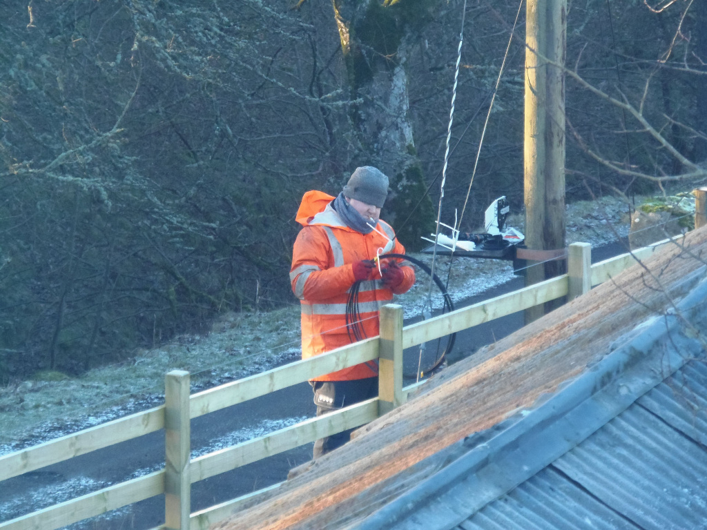 Man, clad in a bright orange high-visibility work jacket over a darker, possibly navy, base layer. His head is covered by a grey knitted beanie. He's positioned slightly off-center, towards the left, working with what appears to be a length of dark fibre optic cable. His hands are gloved, and he's carefully manipulating the cable.  His expression is partially obscured by his beanie and the angle but suggests concentration. The cable is a dark, almost black, and coils slightly as it extends from his hands, suggesting flexibility. It's connected to some small grey or white equipment mounted on a wooden post. This equipment looks technical, possibly a splice case or similar telecoms device. A wooden post-and-rail fence runs diagonally across the mid-ground and foreground, partially obscuring the lower part of the man’s body.