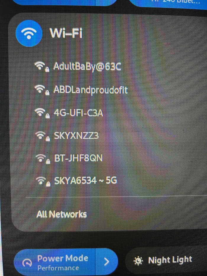 Close-up shot of a digital device screen, specifically showing a Wi-Fi network selection interface. The screen is dark gray/charcoal, providing a stark contrast to the lighter-coloured text and icons. At the top, a stylised Wi-Fi symbol (blue circle with white signal bars) sits to the left of the word Wi-Fi in a sans-serif, slightly bolder typeface. Below this, a list of available Wi-Fi networks is presented, each preceded by a Wi-Fi symbol and a small lock icon indicating password-protected networks. The network names vary in length and character combinations: AdultBaBy@63C, ABDLandproudofit, 4G-UFI-C3A, SKYXNZZ3, BT-JHF8QN, and SKYA6534~5G. At the bottom, an All Networks option is displayed. Further down, two buttons are partially visible: a blue Power Mode button (with a Performance sub-label) featuring a power icon and a right-facing arrow, and a partially visible Night Light button with a sun icon.