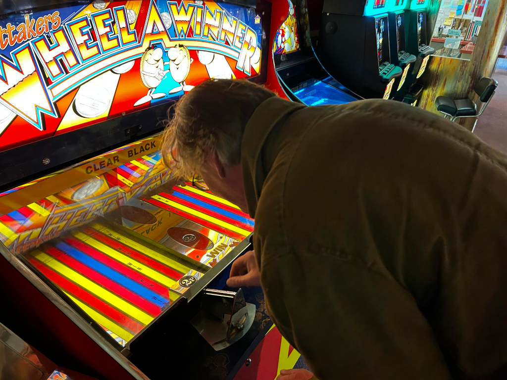 Charlie leaning over a brightly coloured arcade game, his upper body dominating the frame. He's wearing a dark olive-green, possibly corduroy, jacket. His right hand rests near the game's playing area, suggesting interaction with the game. His face is partially visible, angled downwards; only his forehead, eyes (partially obscured), and side of his nose are clearly seen, with focus on the back of his head and his jacket. The game itself is a Wheel-a-Winner style coin pusher, boasting a vibrant colour scheme of bright reds, yellows, blues, and blacks. The game's graphics are cartoonish with lively, rounded figures in a retro style.  The design features a large wheel with many slots to drop coins in. The setting is a dimly lit arcade or game room. Behind the main subject are other arcade games partially visible, contributing a sense of depth. The background features a muted wall with industrial-looking metal components, and a portion of what looks like a wall-mounted shelf or cabinet with some objects on it. 