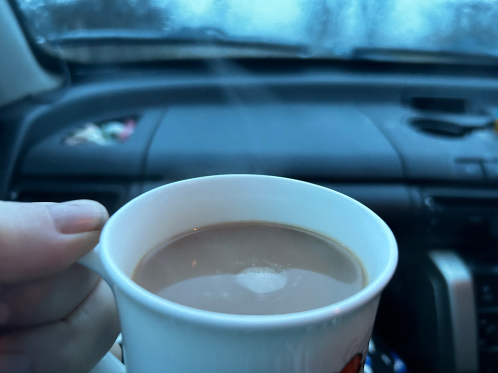 Close-up view of a hand gently cradling a white coffee mug. The mug is slightly angled to the left, revealing a creamy, light-brown liquid inside—presumably coffee or tea—with a subtle foamy texture on its surface. A hint of steam rises from the beverage. The hand holding the mug belongs to a fair-skinned person, only a portion of the hand and fingers are visible. The fingers exhibit a gentle grip, suggesting a comfortable and relaxed state. The mug features a barely visible, slightly blurred design or pattern near its base, hinting at a simple, possibly cartoonish, motif.