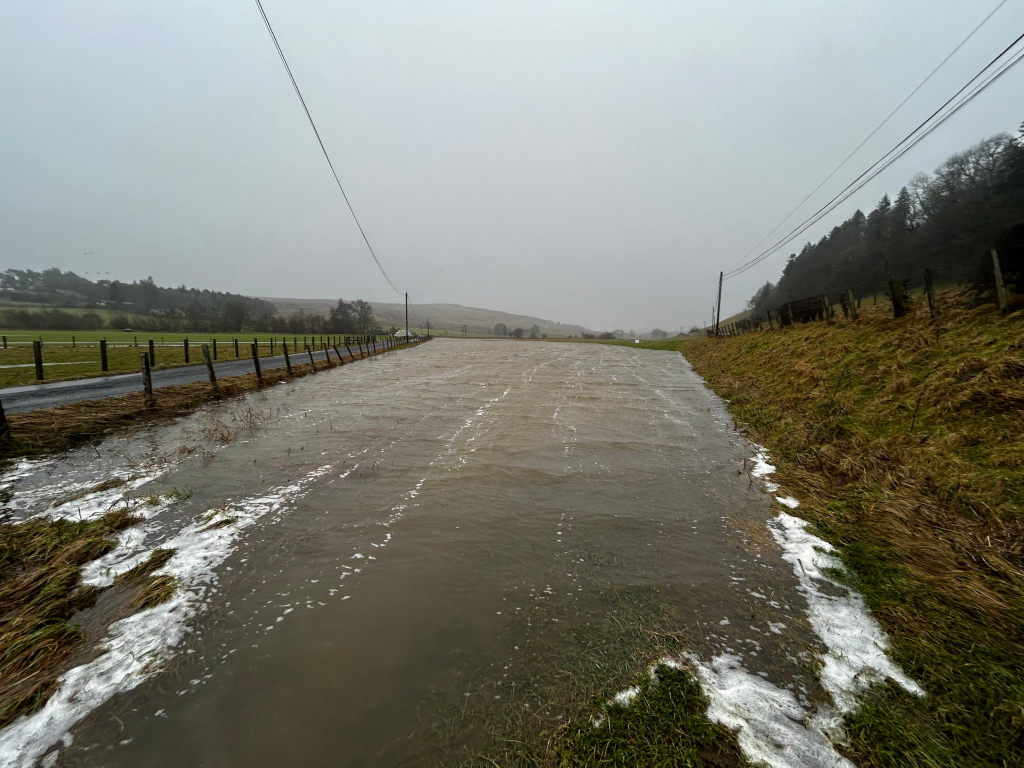 Flooded, narrow waterway or ditch running parallel to a rural road. The water is a murky brown-grey, churned slightly by an unseen current, creating subtle ripples and small, frothy white caps along the edges where it laps against the banks. Along the water's edge, patches of melting snow cling to the grass. The road, visible on the left, is dark grey and appears to be relatively intact. A wooden fence runs alongside the road, acting as a boundary between the roadway and the field. Power lines stretch across the scene, disappearing into the grey distance. The banks of the waterway show various shades of brown and green vegetation, with dry, golden-brown grasses prominent along the right bank.