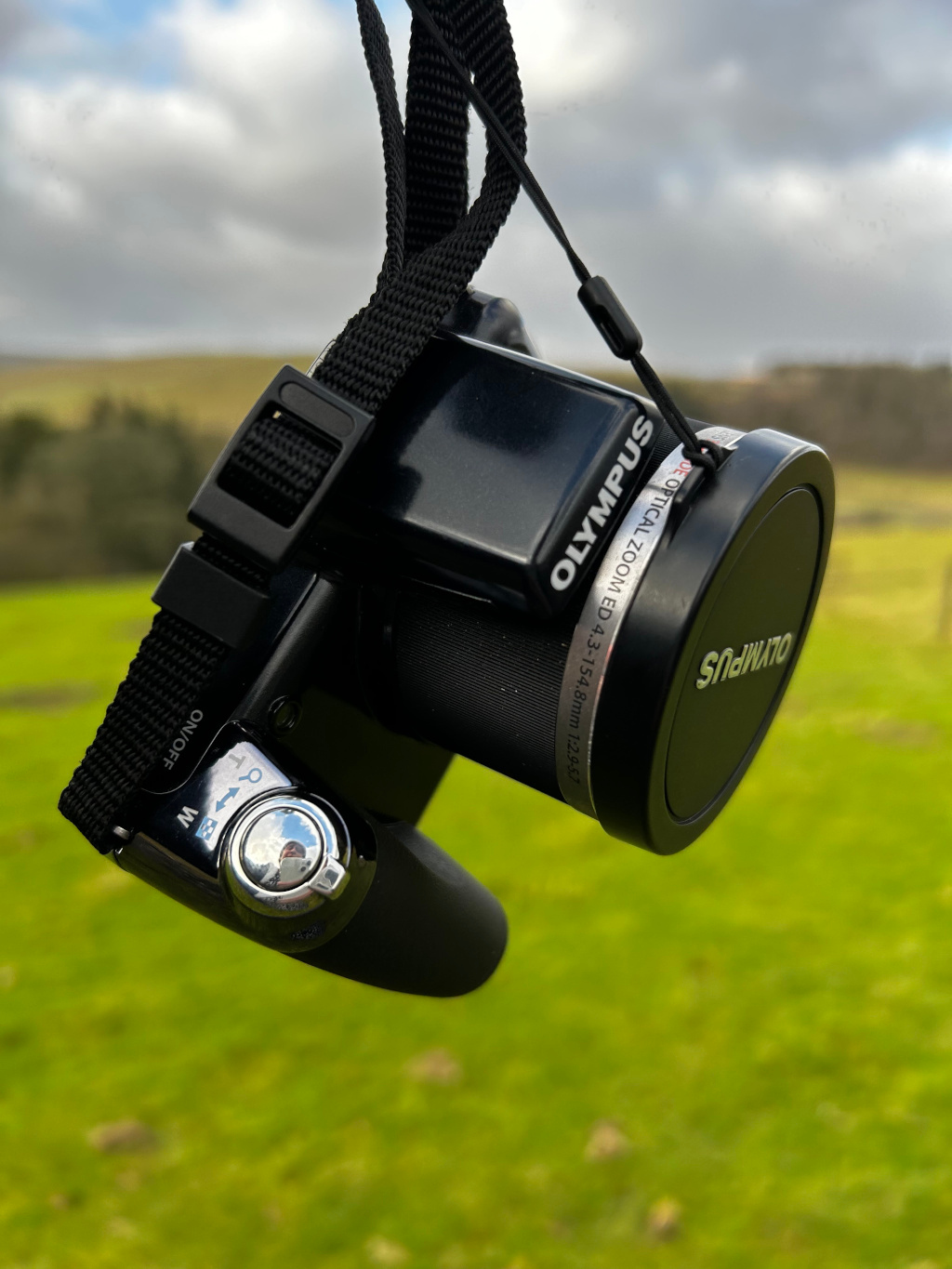 Black Olympus digital camera, hanging upside down from a black neck strap. The camera is compact, with a prominent lens cap that bears the Olympus logo. The lens barrel displays the specifications: OLYMPUS OE OPTICAL ZOOM ED 4.3-154.8mm 1:2.9-5.7. The camera's mode dial is partially visible, showing markings for ON/OFF and W (likely wide-angle mode), indicating a switch or dial for the operating modes. The strap is made of sturdy, woven black material with a clip securing it to the camera. background is a gently sloping landscape, seemingly rural or pastoral. Rolling green hills, dotted with sparse trees and shrubs, stretch into the distance under a cloudy sky. The sky is a blend of light gray, white clouds, and hints of blue, indicating a day with soft natural light, possibly overcast. The green of the fields is vibrant but not intensely saturated, suggesting a soft, possibly cool-toned natural light.