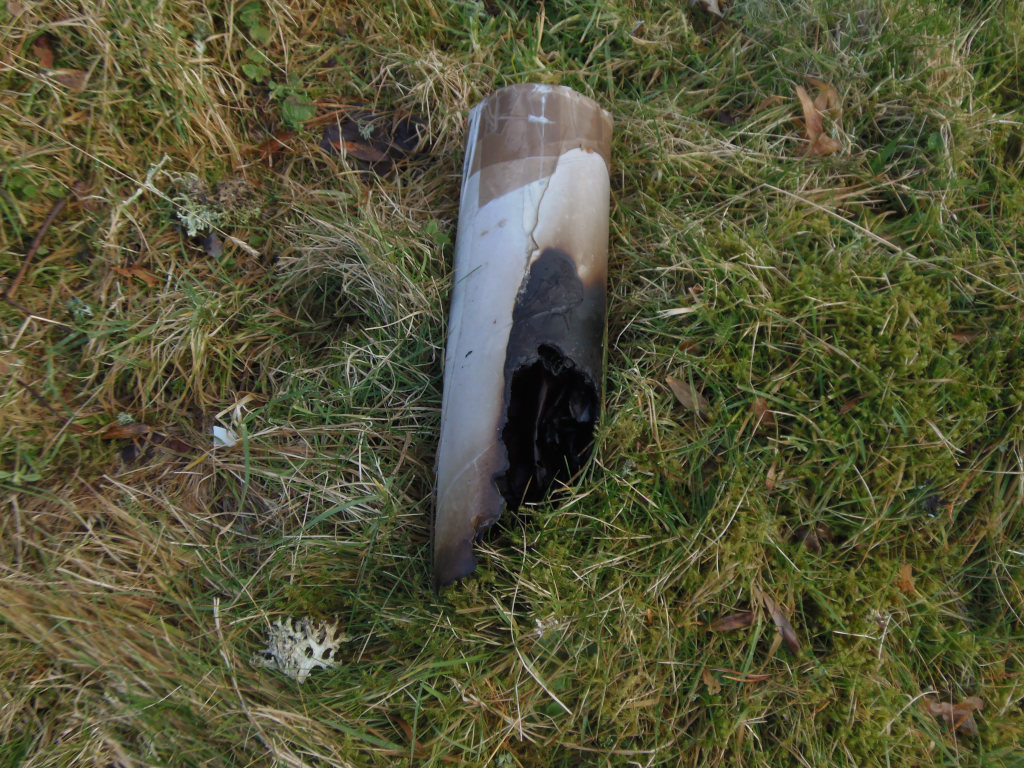 Partially burned cylindrical object, lying on its side in the grass. It appears to be made of a material that's charred black on one side, revealing a lighter, possibly off-white or beige, under layer. The charred portion has irregular, jagged edges, suggesting intense heat. The cylinder is approximately half its original length, the top portion seemingly missing. There is no visible texturing to the unburned portion, but the form suggests it might be cardboard or a similar material. It rests at a slight angle, almost leaning. The scene is set outdoors in a field of short, somewhat sparse grass. The grass is a mixture of muted greens and browns, indicative of a late autumn or early spring setting. Patches of darker green, possibly moss, punctuate the grass. Scattered dried leaves of various browns and muted yellows are visible throughout the grass.