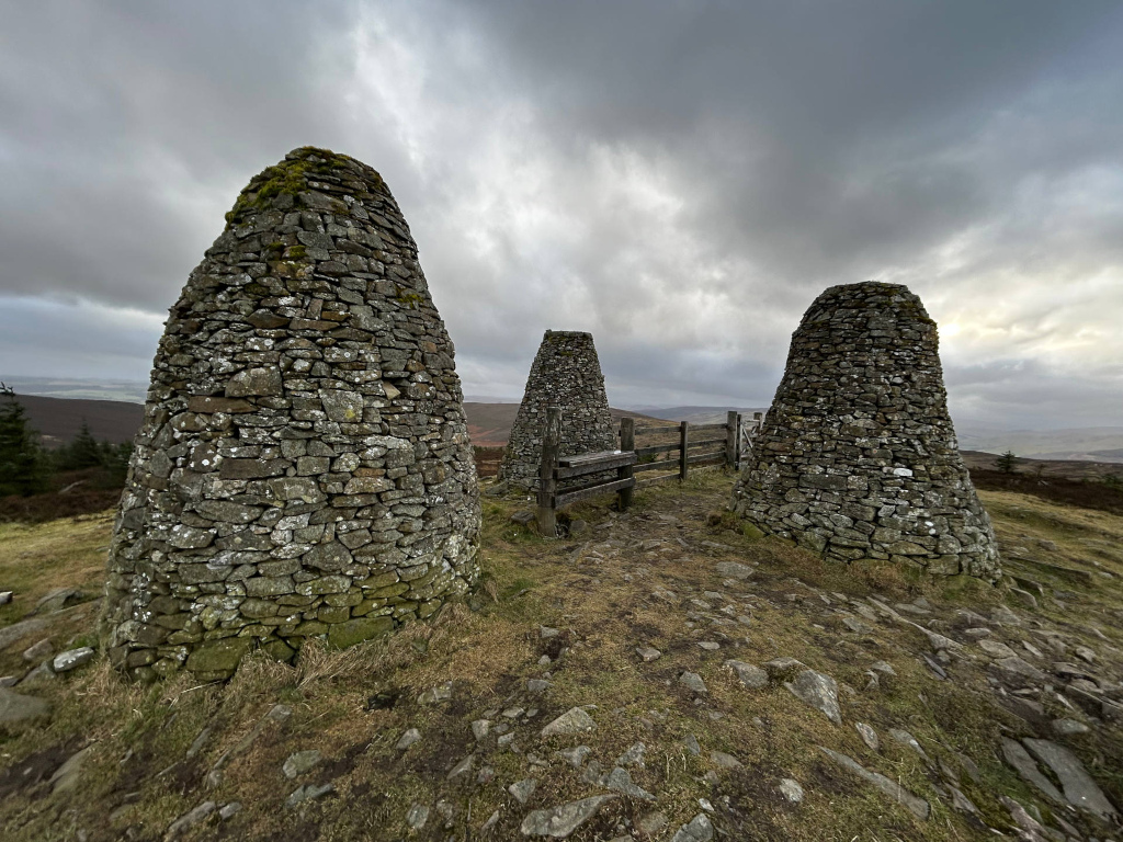 Three conical stone structures, crafted from grey and brown stones of varying sizes, seemingly mortared together. Moss and lichen cling to many stones, especially at the bases, adding texture and age. The structures are roughly the same height but vary slightly in width at their bases. These are clearly ancient, weather-beaten monuments. A simple, dark-brown wooden bench sits between the central and rightmost stone structure, partially obscured by a low, rustic wooden fence or railing. No people are present. The scene is set atop a windswept hill or moorland. The foreground is composed of sparsely vegetated, uneven ground with exposed patches of dark soil and small rocks scattered across the short, dry grass. The background showcases a hazy, expansive landscape under a dramatically overcast sky. The sky dominates the upper two-thirds of the image, a brooding blend of dark greys and whites, suggesting an impending storm or recently passed one. Distant, faint, rolling hills are visible under the clouds, giving a sense of depth.