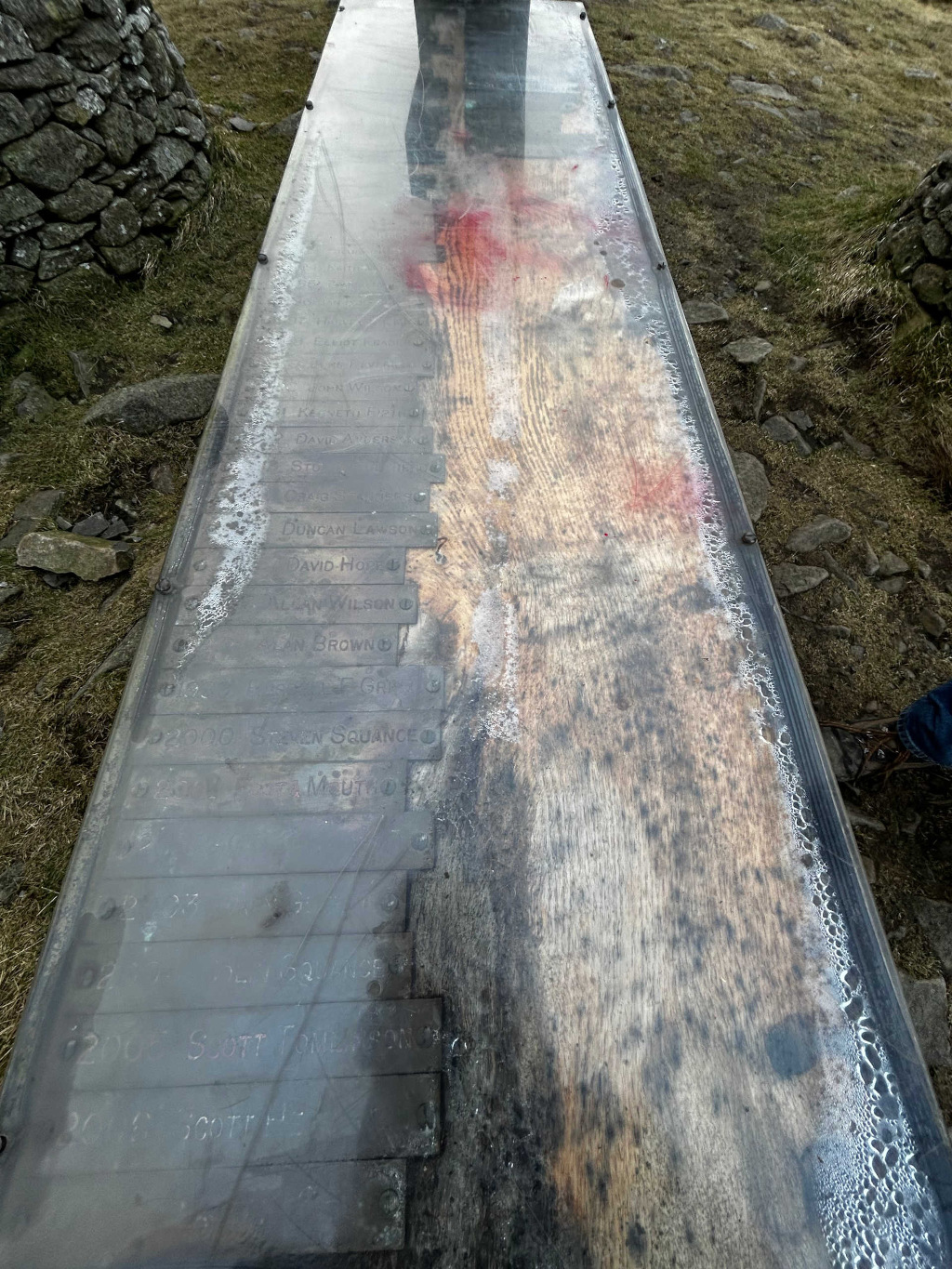 Long, rectangular slab of weathered wood, seemingly embedded within a clear, protective acrylic or glass casing. This casing is slightly elevated from the ground, acting as a monument or memorial. The wood displays a rich tapestry of natural grain, heavily marked by time and the elements – a deep, smoky grey-brown hue with streaks of lighter, almost bleached wood. Embedded into the wood are numerous rectangular metal plaques, each bearing a name and sometimes a number (partially legible due to weathering and possibly frost). These plaques are uniformly spaced and aligned, creating a sense of ordered record-keeping despite the organic texture of the wood. A reddish stain or discoloration is prominently visible along a vertical section of the wood, adding a mysterious contrast to the otherwise subdued palette. Water droplets or condensation are visible along the edges of the protective casing, suggesting exposure to cold, damp conditions. A portion of a person's leg and foot, clad in jeans and brown boots, is visible in the bottom right corner, implying a viewer or passer by present at the site.