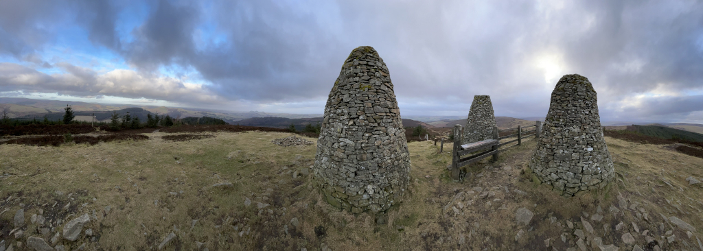 Three prominent, conical stone structures, seemingly cairns or small towers, built from rough, grey and brown stones of varying sizes. They are positioned on a gently sloping, grassy hilltop.  The largest cairn is positioned slightly left of center, and the other two are smaller and situated to its right, creating a somewhat triangular arrangement. Between the two smaller cairns sits a simple, rustic wooden bench, seemingly built into a low stone fence that partially encircles the three structures. No people are present. The scene unfolds on a windswept moorland or upland area. The foreground is a mix of short, dry grass, patches of bare earth, and scattered rocks, typical of a high-altitude landscape. The background features a panoramic view of rolling hills extending to the horizon, covered in a mixture of heather and dark green coniferous trees.  The sky dominates the upper third of the image, filled with a dramatic, cloudy expanse. The clouds are mostly grey and white, with some darker, brooding areas, creating a sense of depth and movement. A hint of lighter blue sky peeks through in places, and a soft, diffused light suggests either early morning or late afternoon.