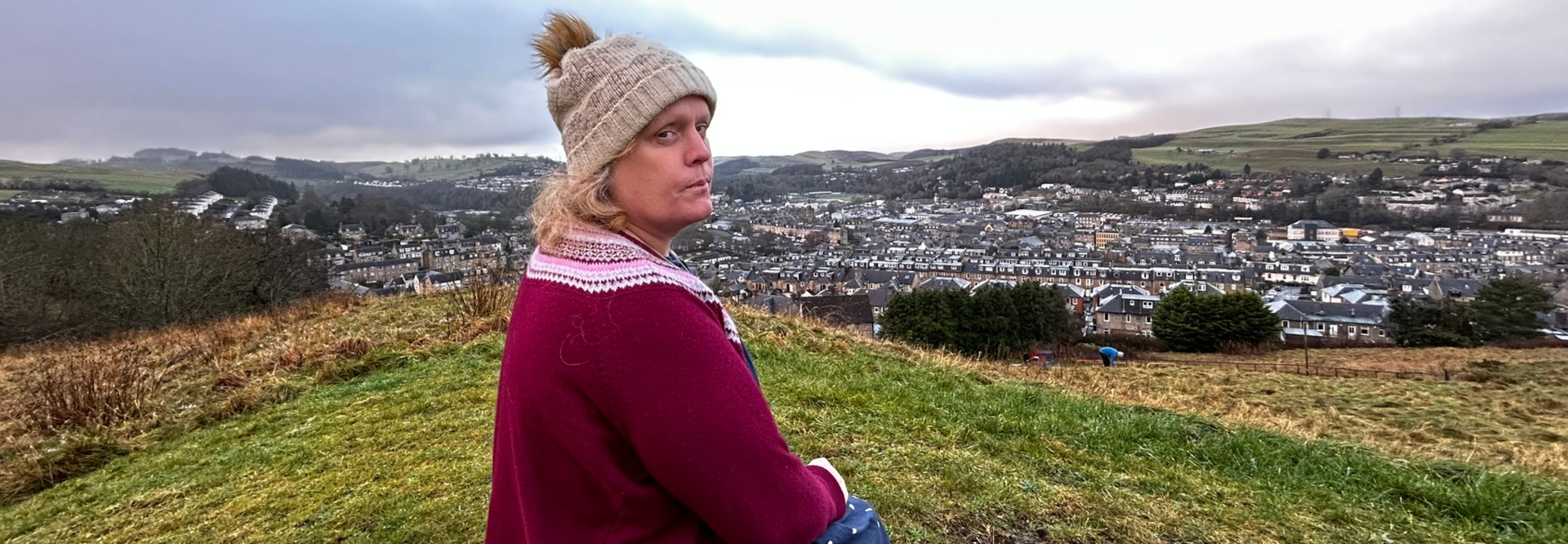 Middle-aged woman (Leonie), her face slightly turned away from the viewer, positioned three-quarters to the right. She’s wearing a chunky, oatmeal-coloured knit beanie with a fluffy pom-pom atop, framing a face with a soft, round visage. Her expression is pensive, almost melancholic, with a slightly downturned mouth. She’s clad in a deep burgundy, possibly wool, sweater with a subtle, Fair Isle-style patterned yoke in pale pink and white. The sweater's texture appears soft and thick. A hint of a patterned, possibly denim, fabric is visible at her lower hem. Her blonde hair peeks out from under the beanie, framing her face. Her hands are partially visible, resting near her abdomen. She appears to be seated or perched on a grassy incline. The scene is an expansive vista, dominated by a sprawling town nestled in a valley. The town is a collection of tightly packed, predominantly grey-roofed houses, appearing in muted shades of grey, stone, and muted browns. The background reveals rolling green hills, softened by a light overcast sky that is predominantly a pale grey-blue, suggesting an early morning or late afternoon, with muted lighting conditions. There's a subtle hint of faint bluish-grey shadows indicating diffuse light, not harsh direct sunlight. The foreground is a gently sloping grassy hill, with a variety of browns and greens in the grasses; shades of yellowish-brown appear particularly toward the lower left. This hillside contrasts the vibrant colour of her burgundy sweater. A few distant trees dot the landscape. A small figure, seemingly a person, can be seen in the distance, near the town.