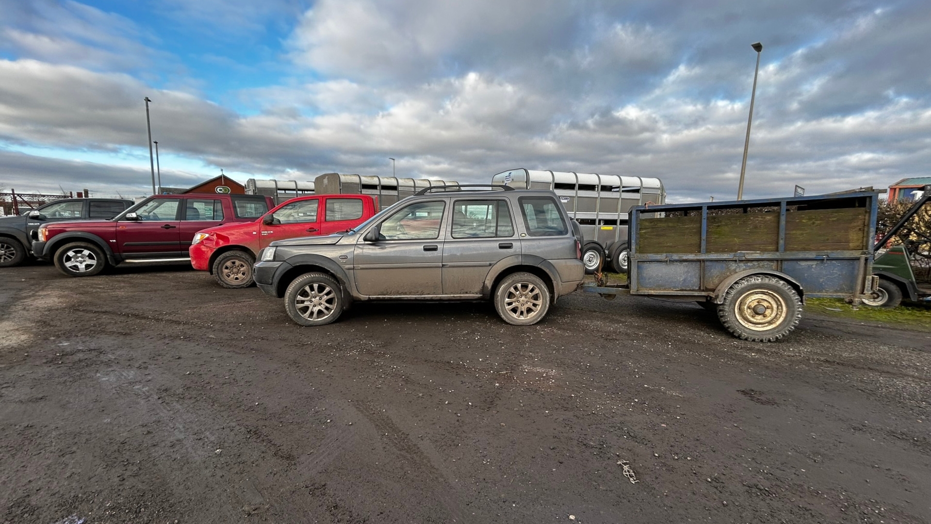 Muddy, gray Land Rover Freelander parked in a gravel lot. It's angled slightly to the right, pulling a weathered, blue single-axle trailer with wooden sides. The trailer appears old and worn, with visible age and dirt. Behind the Land Rover are several other vehicles: a dark red Land Rover Discovery, a red pickup truck (possibly a Toyota Hilux), and a dark-coloured SUV (partially obscured). In the far background, there are several horse trailers, typical of a farm or equestrian centre. The vehicles are all stationary and parked. No people are visible.