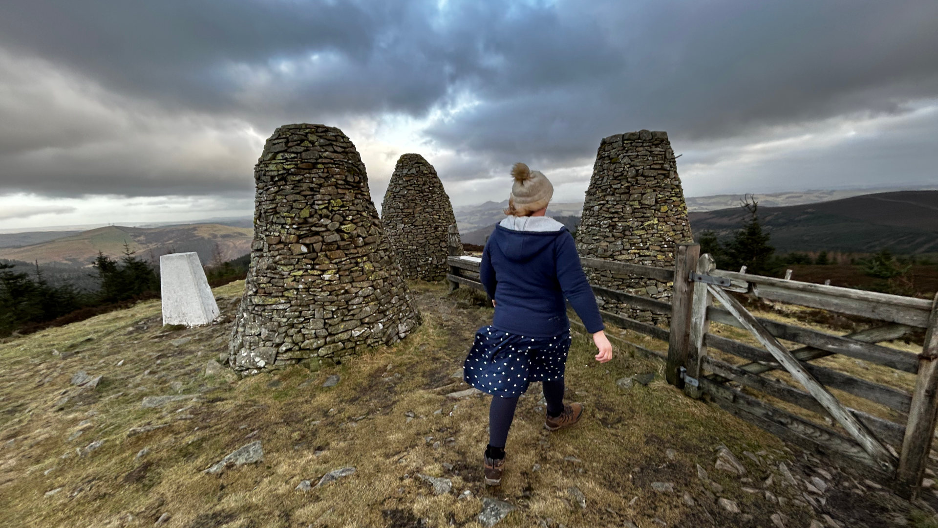 Leonie walking away from the camera towards three stone cairns on a hilltop. The sky is overcast and dramatic; a wooden fence is visible to the right. The overall scene evokes a sense of solitude and exploration in a wild, open landscape. A small, white trig point is also visible near the base of one of the cairns.