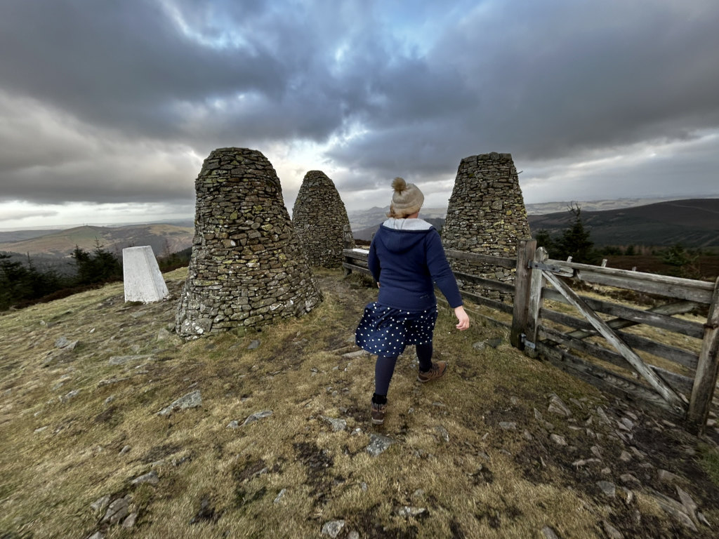 Leonie walking away from the camera towards three stone cairns on a hilltop. The sky is overcast and dramatic; a wooden fence is visible to the right. The overall scene evokes a sense of solitude and exploration in a wild, open landscape. A small, white trig point is also visible near the base of one of the cairns.