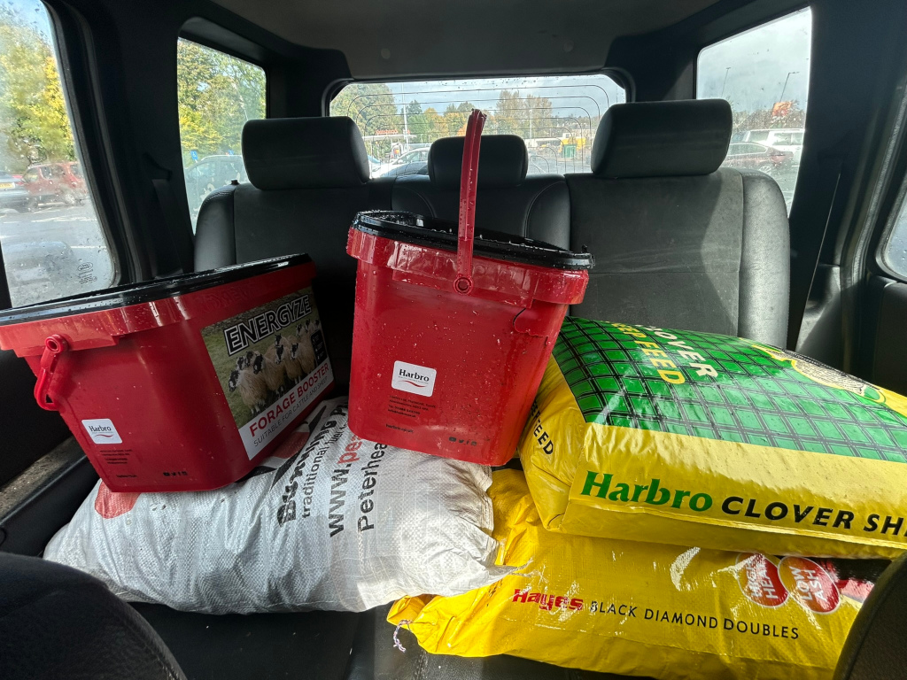Backseat of a vehicle filled with livestock feed. There are two red plastic buckets, one containing EnerGize Forage Booster (Harbro brand), the other is partially filled. Stacked on the rear seat are several bags of Harbro brand feed, including clover sheep feed and Black Diamond Doubles. The overall impression is of a farmer or agricultural worker transporting supplies for their animals. The image is straightforward, lacking artistic elements; its primary purpose appears to be documentation.