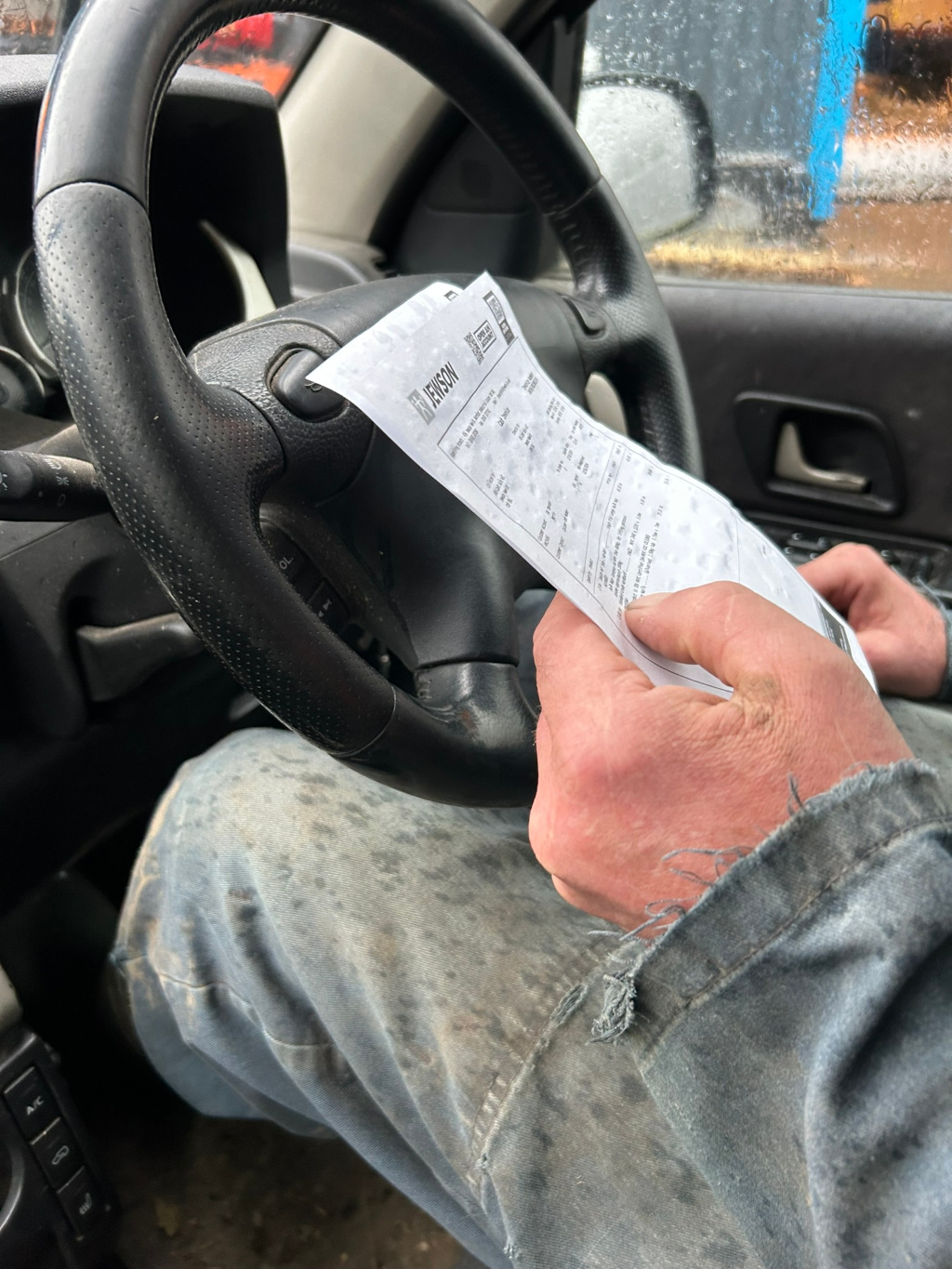 Close-up view of a Charlie's hands holding a receipt or invoice from a company called Jenson. Charlie is seated in the driver's seat of a vehicle, their hands and a portion of their work-worn denim clothing visible. The car's steering wheel is partially in view, suggesting a moment of pause or review of the document. The overall impression is one of a working-class individual reviewing a business transaction.
