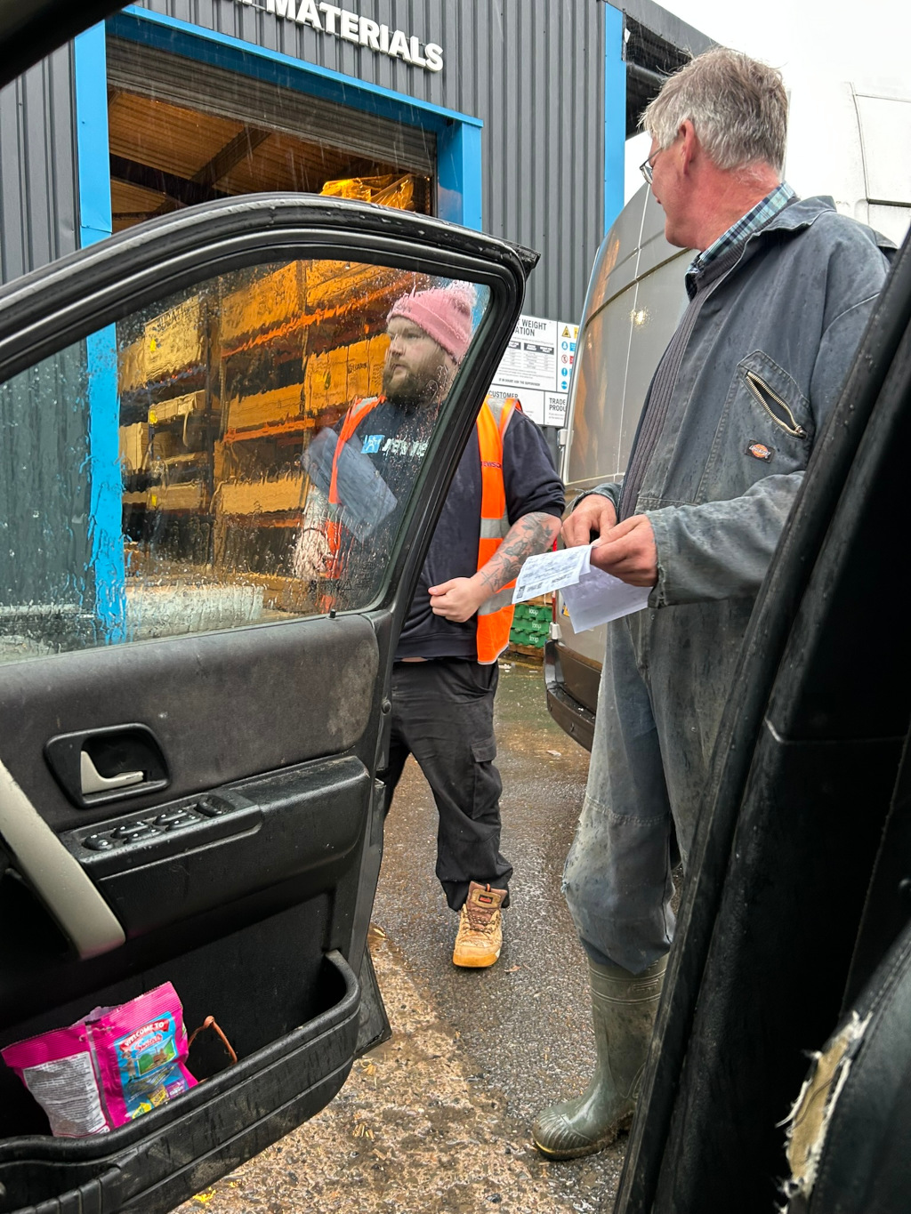 Two men outside a building labelled MATERIALS. One man, a shop worker is wearing a pink beanie and an orange safety vest, is standing near a vehicle. He appears to be handing something to the other man, who is older and dressed in work overalls and green rubber boots. Charlie is holding a piece of paper, possibly a receipt or invoice, suggesting a transaction or delivery is taking place. The scene is set in what seems to be a building material supply yard on a rainy day, with rain visible on the car windows. A bag of what looks like pet food is visible in the car's door pocket. The overall mood is one of a casual business interaction.