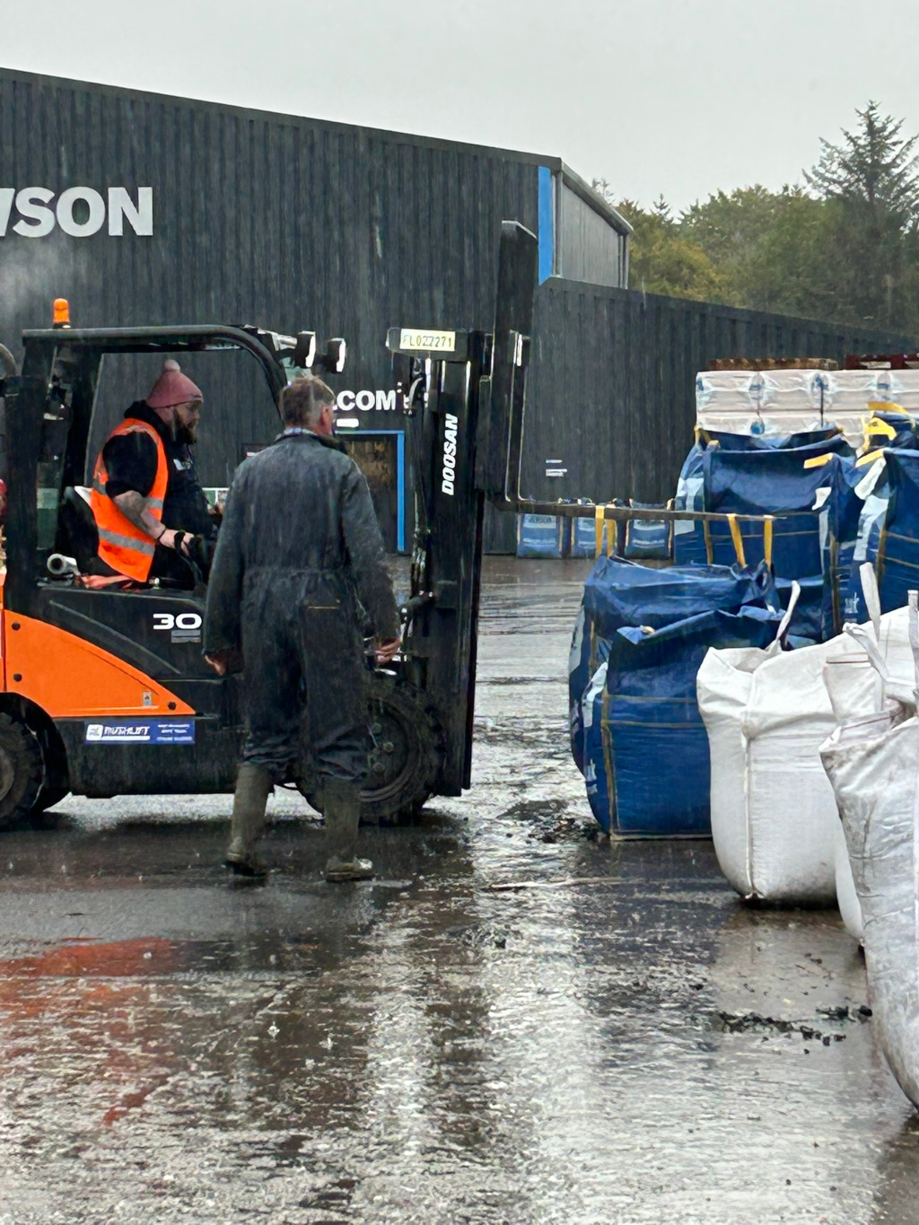 Rainy day scene at what appears to be a construction or industrial site. Charlie in a dark overall is standing near an orange forklift, seemingly overseeing the unloading of large, stacked bags (some blue, some white/off-white) from the forklift. Another man in a high-visibility vest is seated in the forklift. The background features a dark-grey building with SON visible on the side, indicating a company name. The overall impression is one of outdoor manual labour in inclement weather.