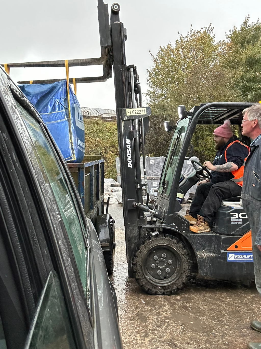 Forklift in a construction or industrial setting. A shop worker is operating the forklift, which appears to be lifting a large, blue tarp-covered object from a small trailer and positioning it near a car. Charlie is standing nearby, observing the process. The scene is outdoors, on a wet, muddy ground. The overall impression is of a routine materials-handling task.