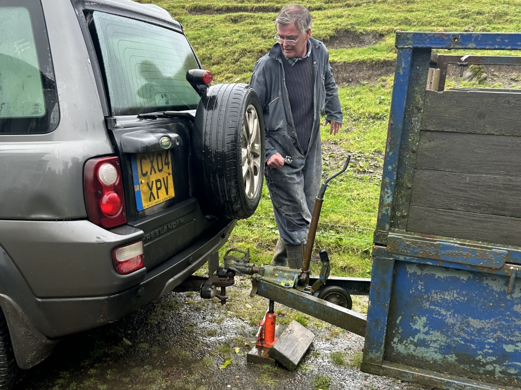 Charlie in work overalls attaching a small, rusty blue trailer to the tow hitch of a grey Land Rover Freelander. The man appears to be using a jack to assist with the connection. The setting is a rural, grassy area.
